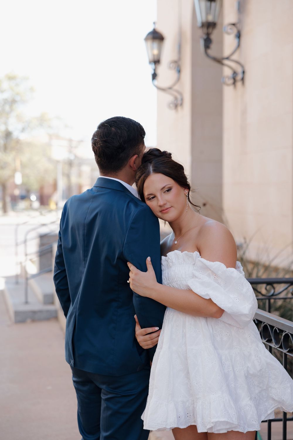 Elegant couple embracing outdoors, woman in white off-shoulder dress looks back while man in navy suit kisses her cheek.