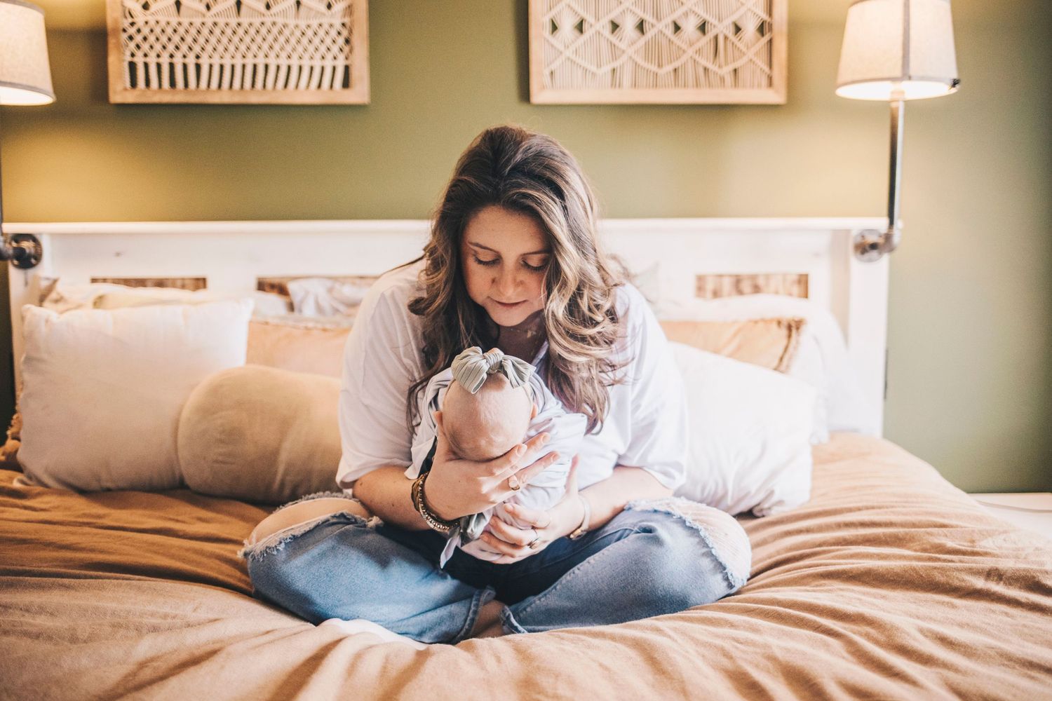 New mother sitting on bed cradling sleeping newborn baby in cozy bedroom.