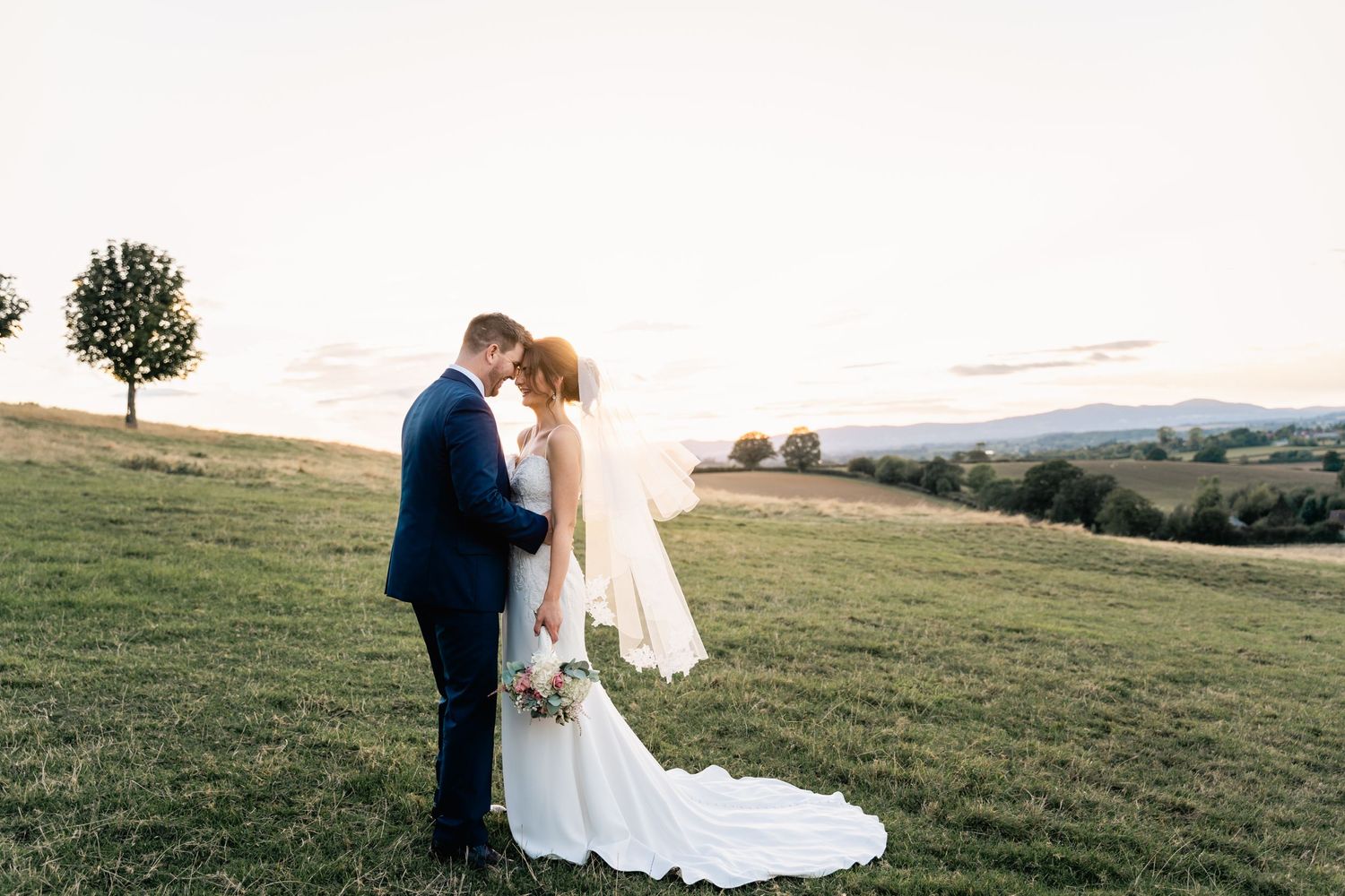 A couple shares an intimate moment on a Worcestershire hilltop overlooking a scenic mountain landscape at sunset.