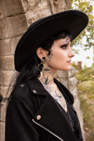 Side profile portrait with wide black hat and neck tattoos against stone wall.
