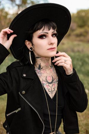 Gothic model in black leather jacket and wide brim hat showing neck tattoos and earrings against natural backdrop.
