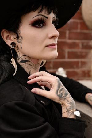 Close up portrait showing black wide brim hat, neck tattoos and bone earrings against brick wall.