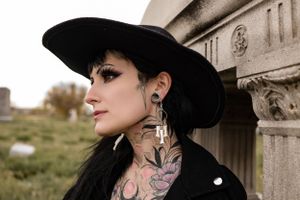 Dramatic profile shot featuring wide-brimmed hat and floral neck tattoos against architectural backdrop.