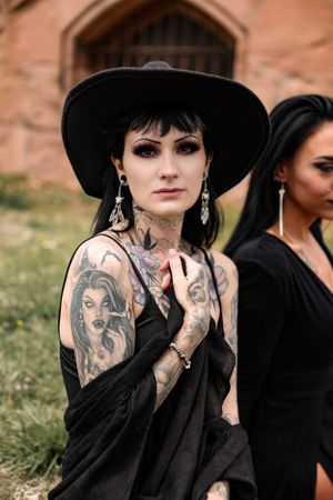 Alternative fashion portrait featuring black hat, neck tattoos and flowing black clothing outdoors.