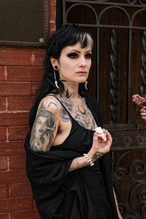 Gothic styled portrait against brick wall showcasing neck tattoos and black clothing.