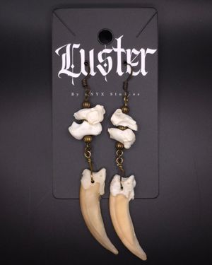 Gothic style earrings with white bone fragments and animal tooth pendants on black display card.