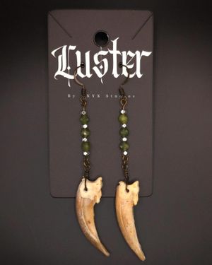 Dangling earrings with carved bone fangs and green crystal beads on a dark Luster display card.