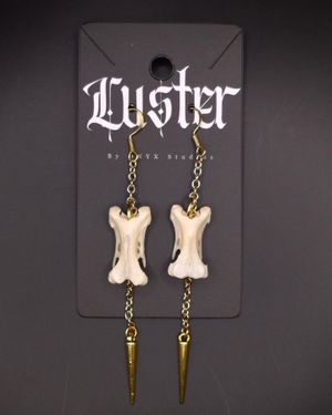 Bone-shaped earrings with metallic spike pendants on brass chains displayed on dark backdrop.