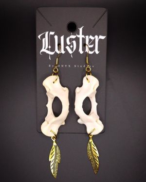 Bone-inspired earrings with brass feather charms displayed on dark Luster branded card.