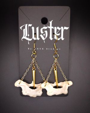 White bone-shaped earrings with brass spikes suspended on chains displayed on a black Luster card.