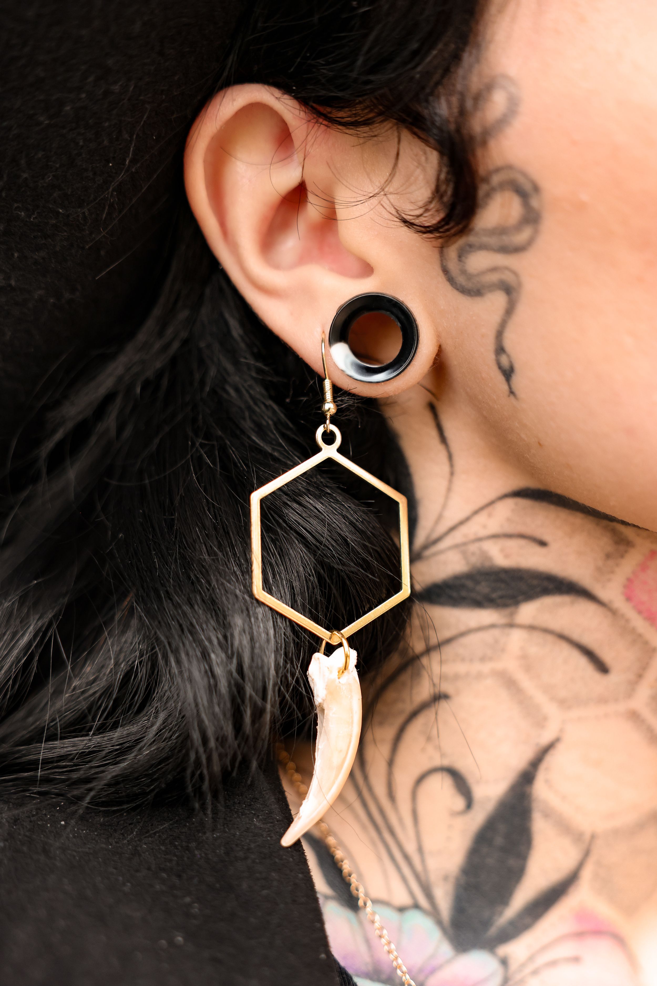 Close up of hexagonal bone earring and neck tattoo detail against black hair.