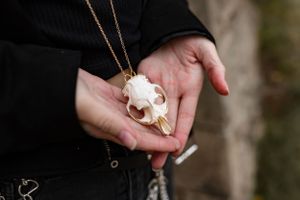 Hands holding small white animal skull necklace pendant against dark clothing.