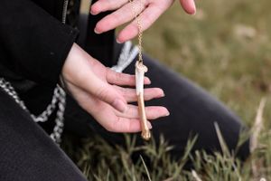 Hand holding bone-shaped pendant necklace against black clothing and grass background.