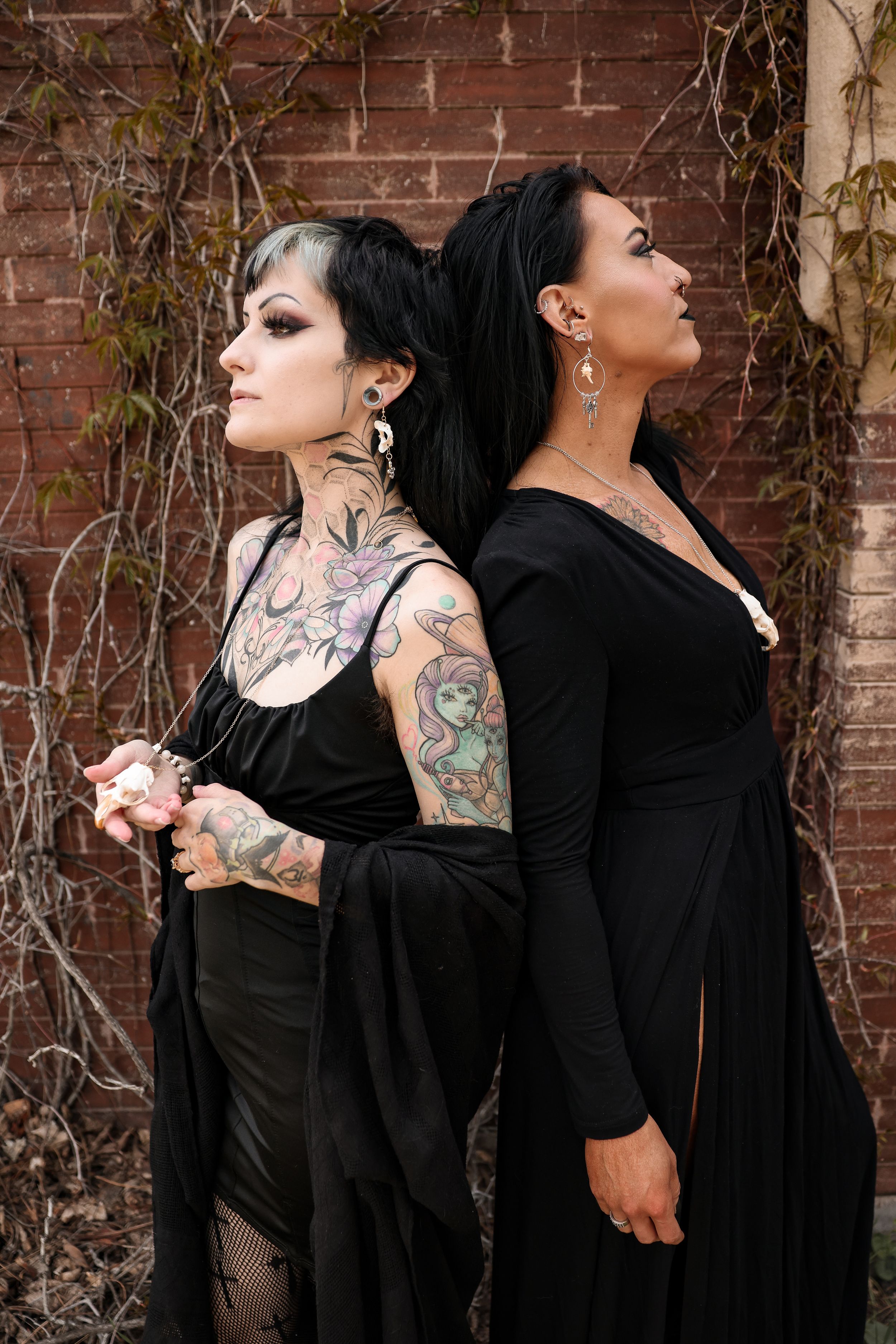 Two models in black dresses pose against brick wall with climbing vines.