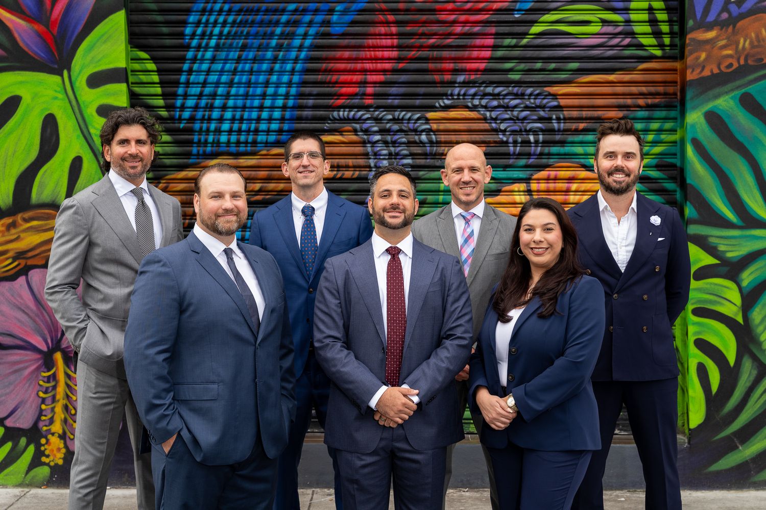 Seven business professionals in formal attire pose together smiling in front of a vibrant colorful mural.