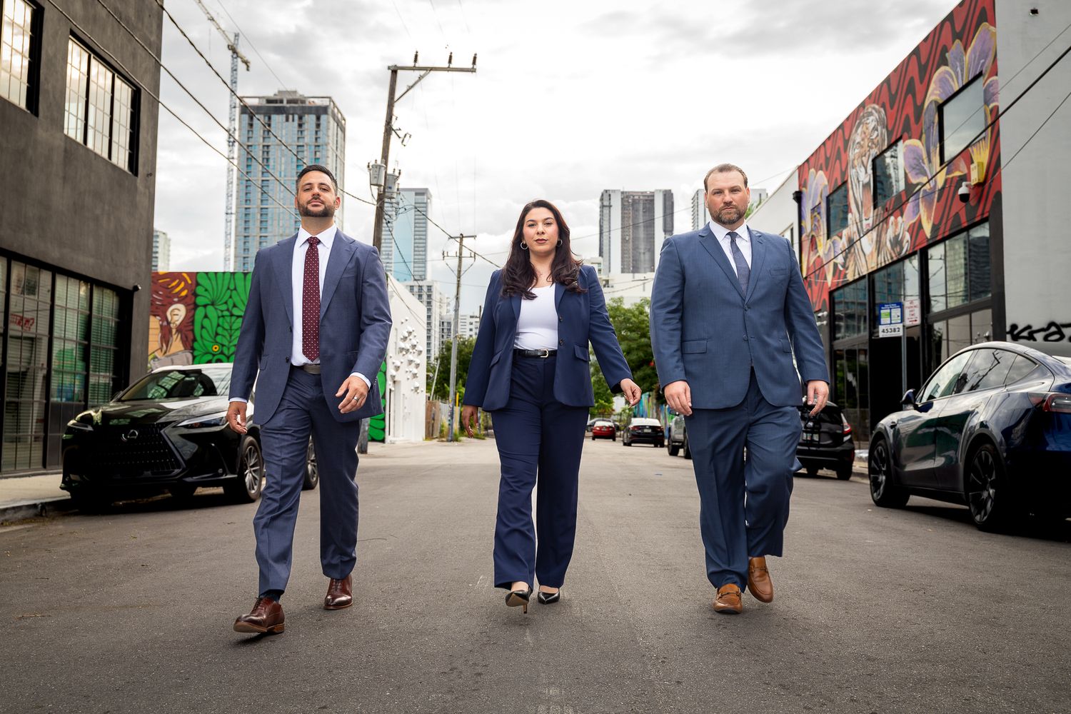 Three business professionals in suits walking confidently down an urban alley with colorful murals and city skyline.
