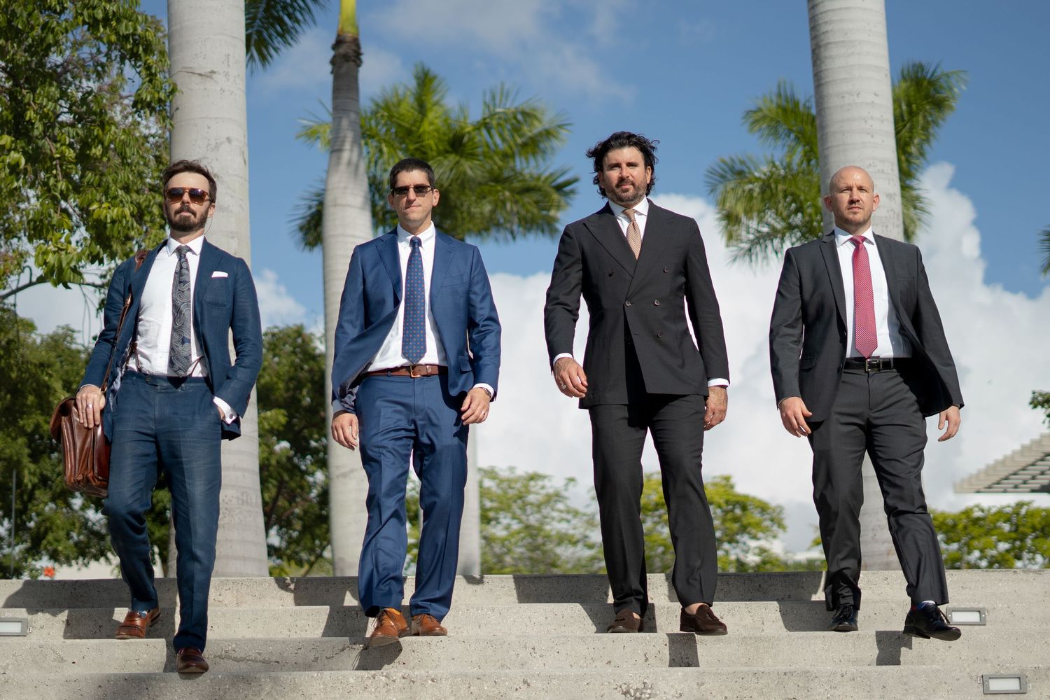 Four men in business suits walking confidently down stairs with palm trees in background.