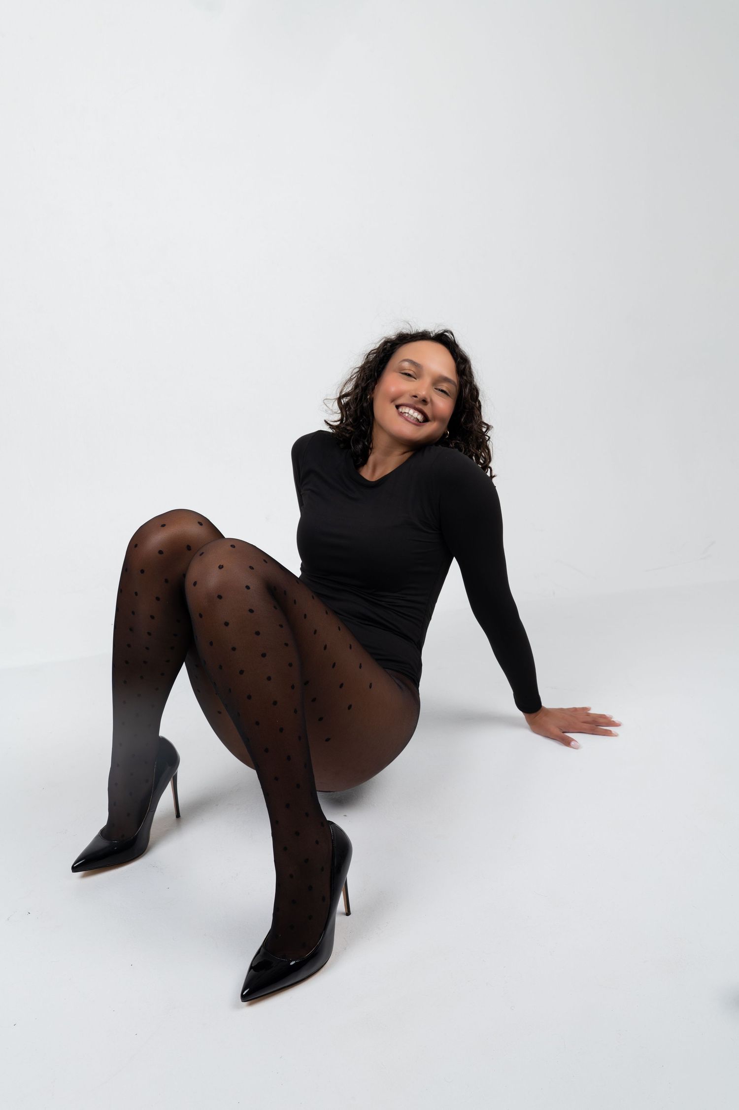 Woman in black bodysuit and polka dot tights sitting on white floor, smiling, wearing black patent heels.