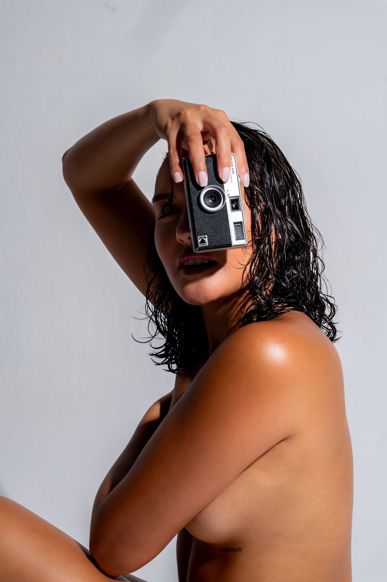 Woman with wet curly hair holding a vintage film camera in front of her face against a light gray background.