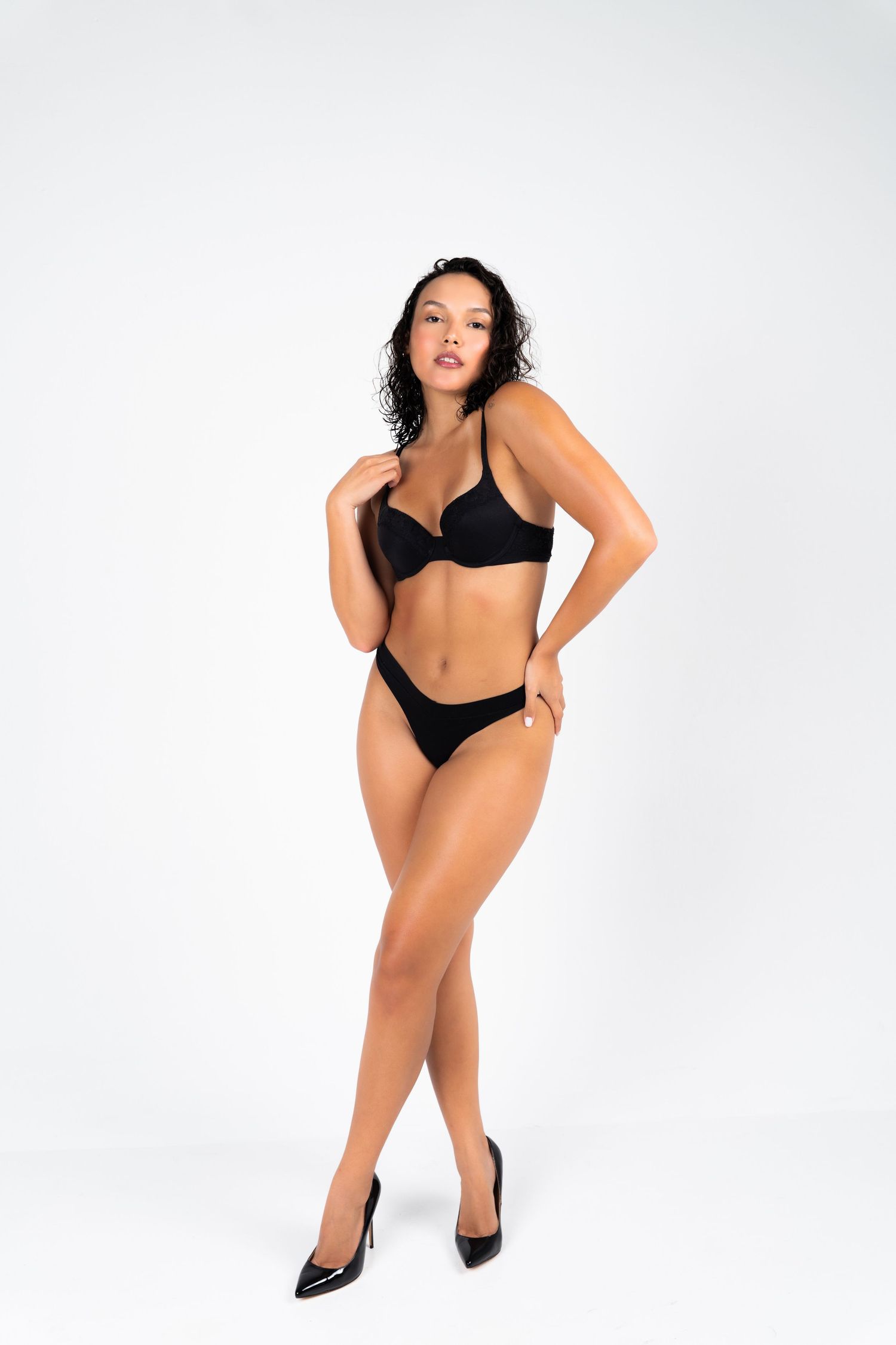 Woman in black lingerie set with heels posing confidently against a white studio background.