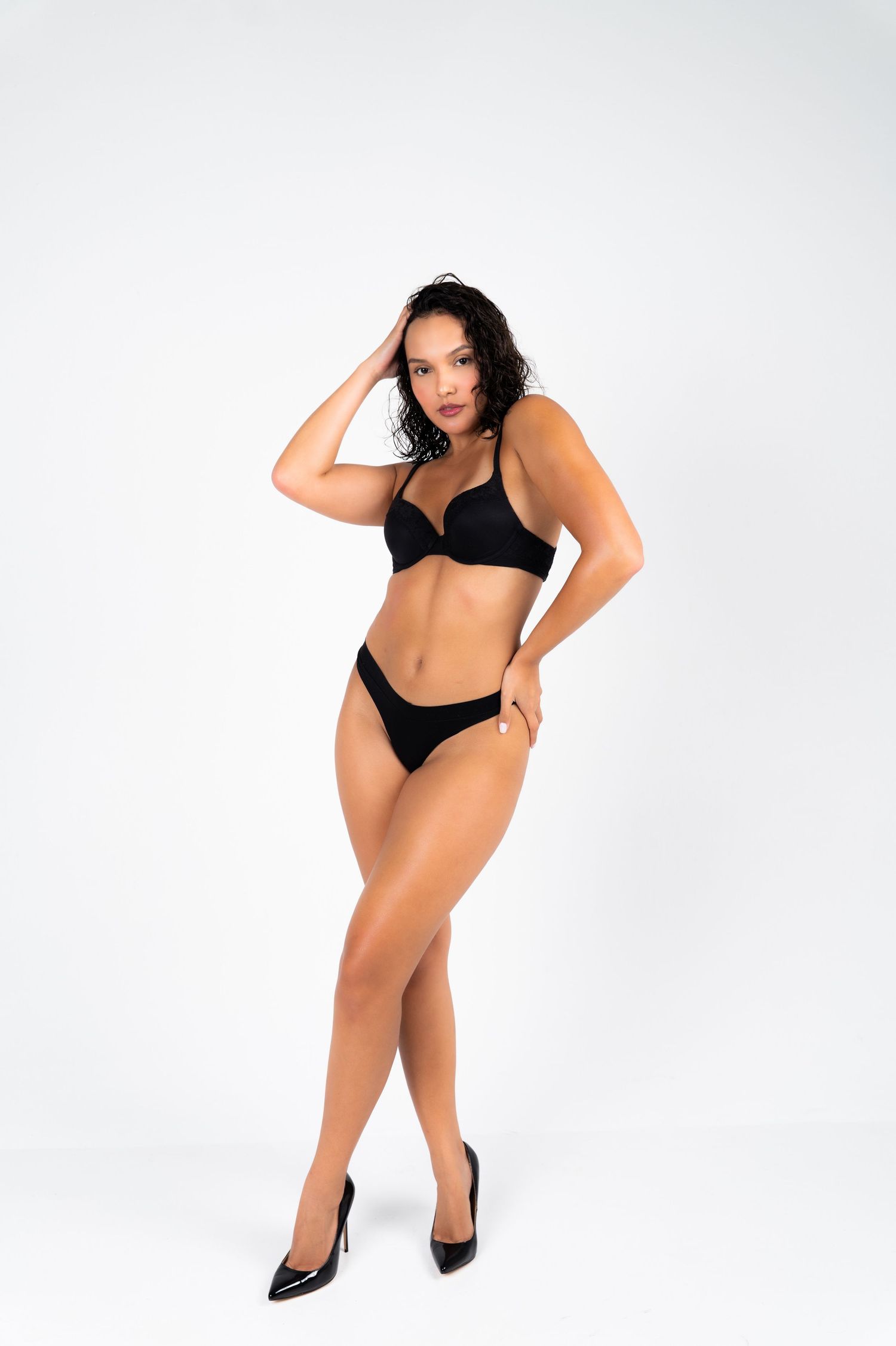 Woman in black lingerie set and heels posing against white background, showcasing fitted bra and bikini bottoms.