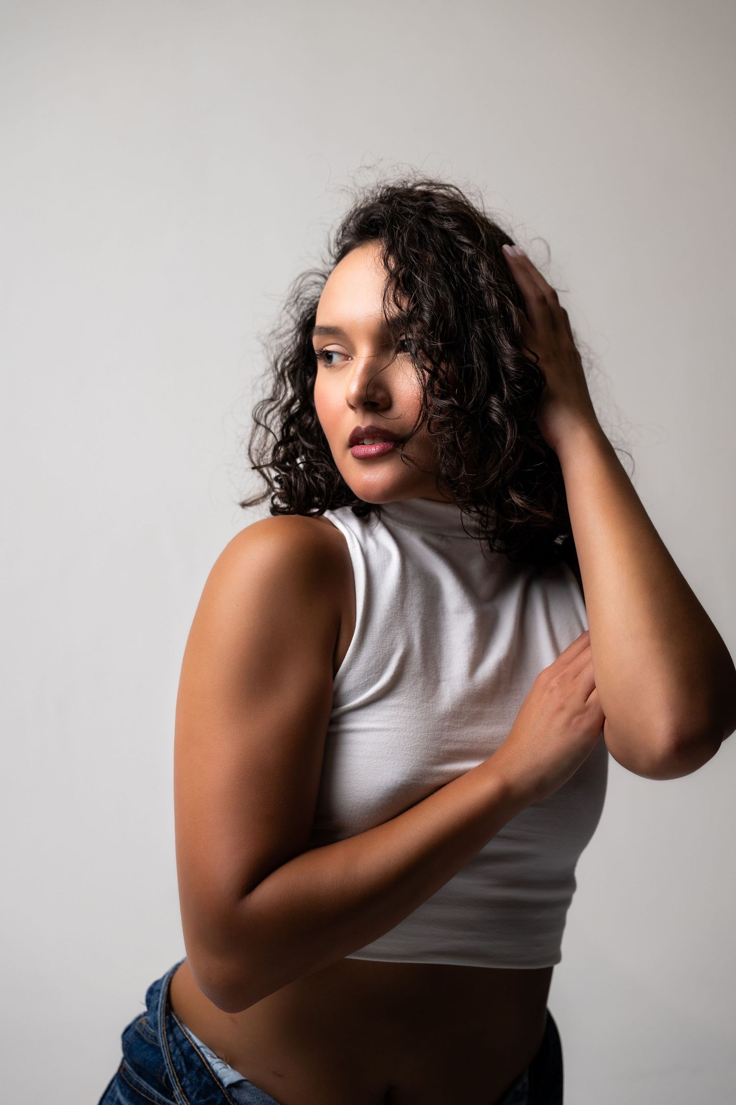 Young woman with curly hair in white crop top and jeans posing confidently against a light gray background.