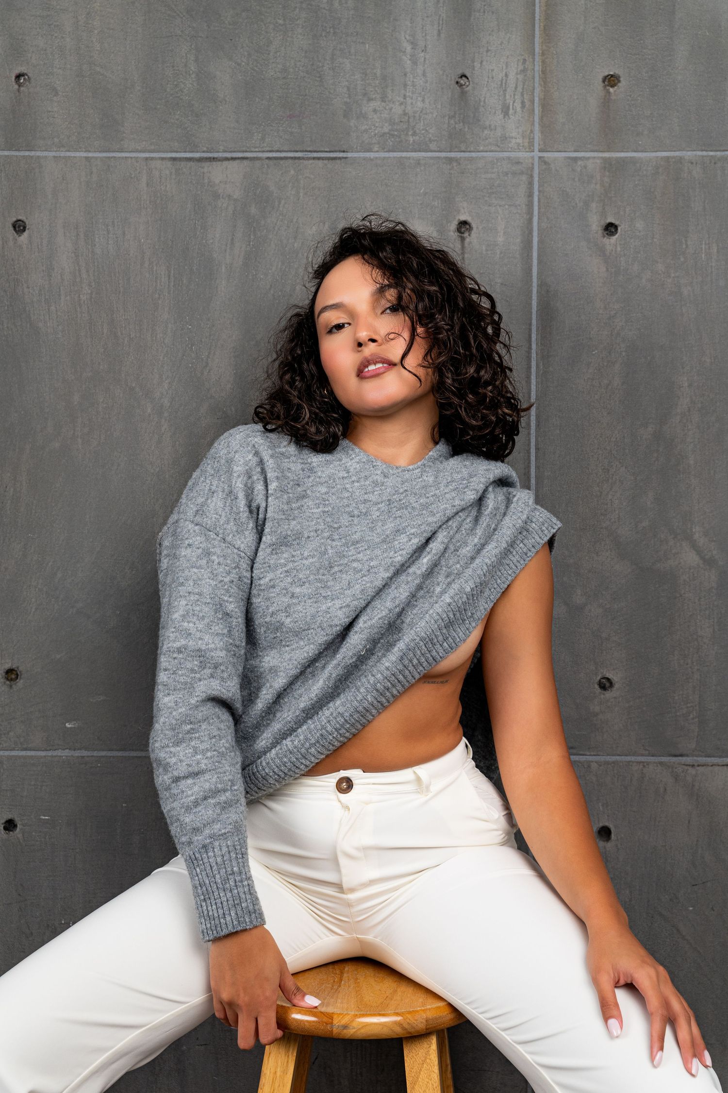 Woman in gray asymmetric sweater and white jeans sitting on wooden stool against concrete wall.
