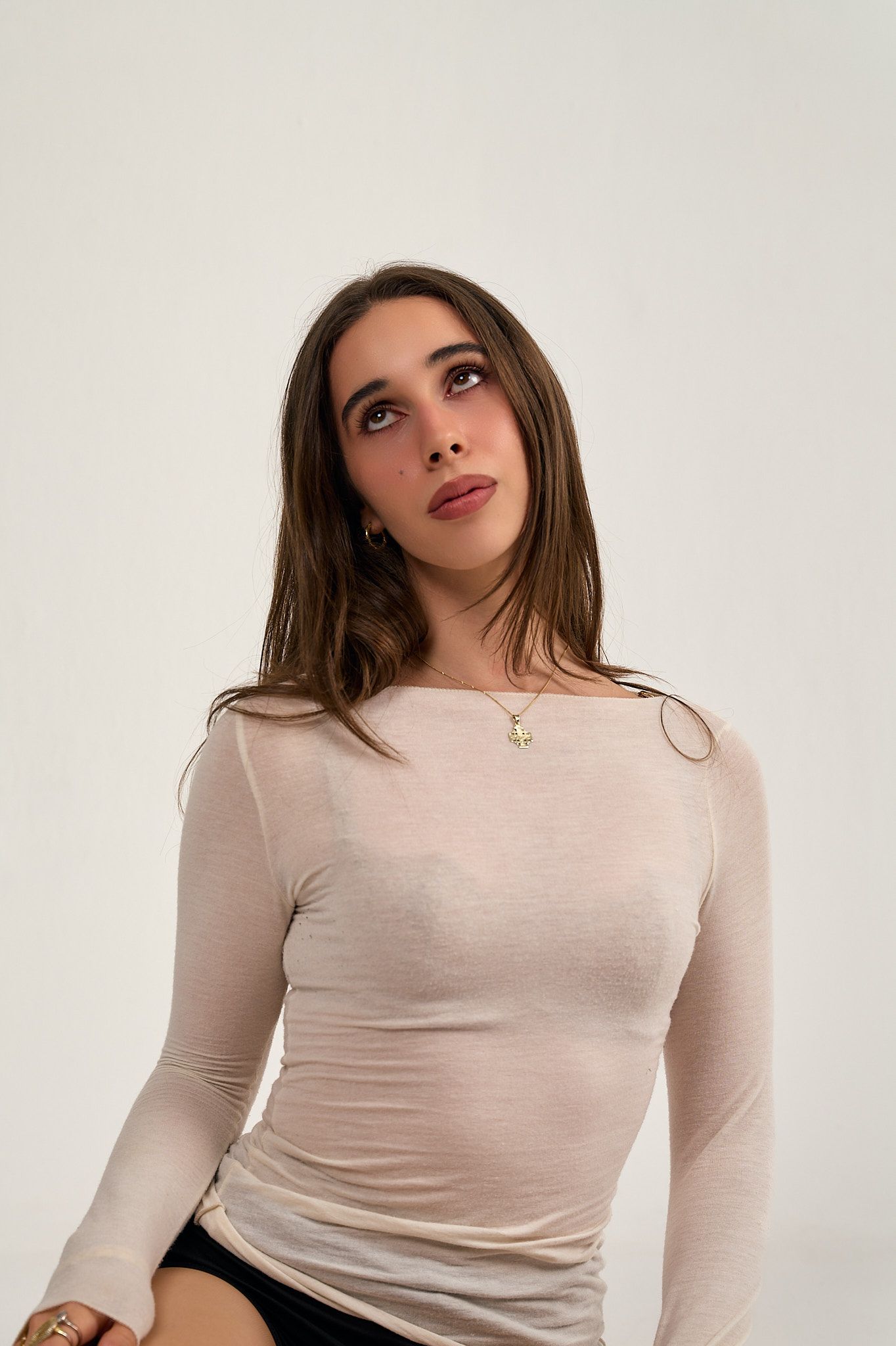 Young woman in sheer beige long-sleeve top with gold necklace posing against white background.