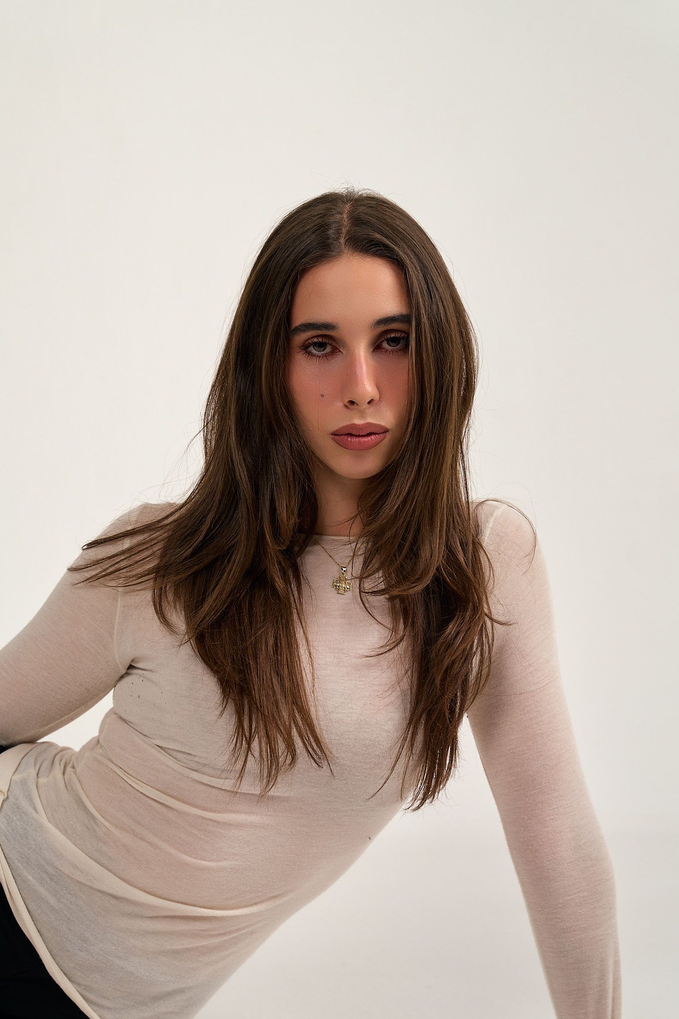Young woman with long brown hair wearing a beige long-sleeve top, posing against a white background.