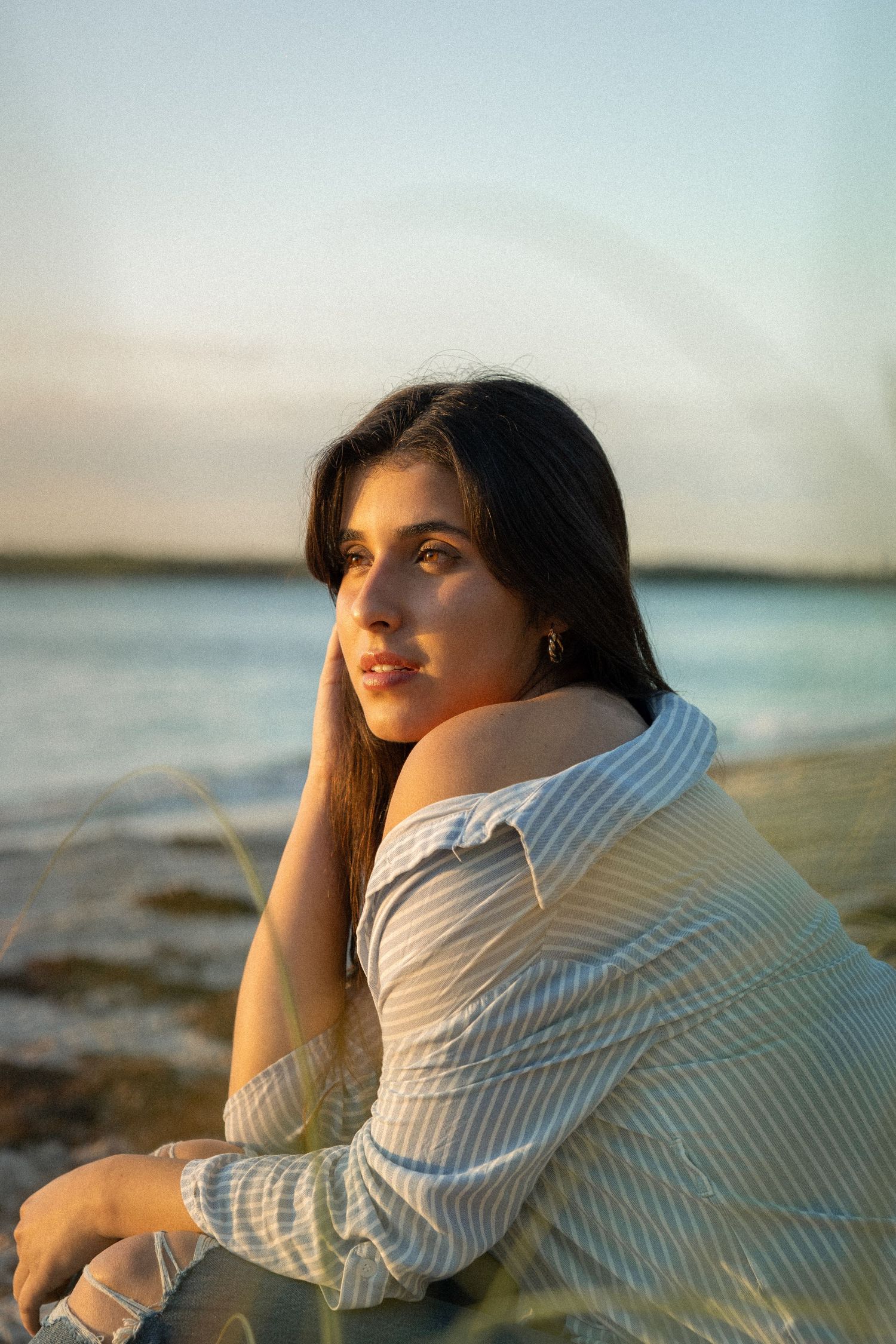 Sabrina Palma. Woman in a white sweater sits on a beach at sunset, gazing upward with long dark hair flowing in the golden light.