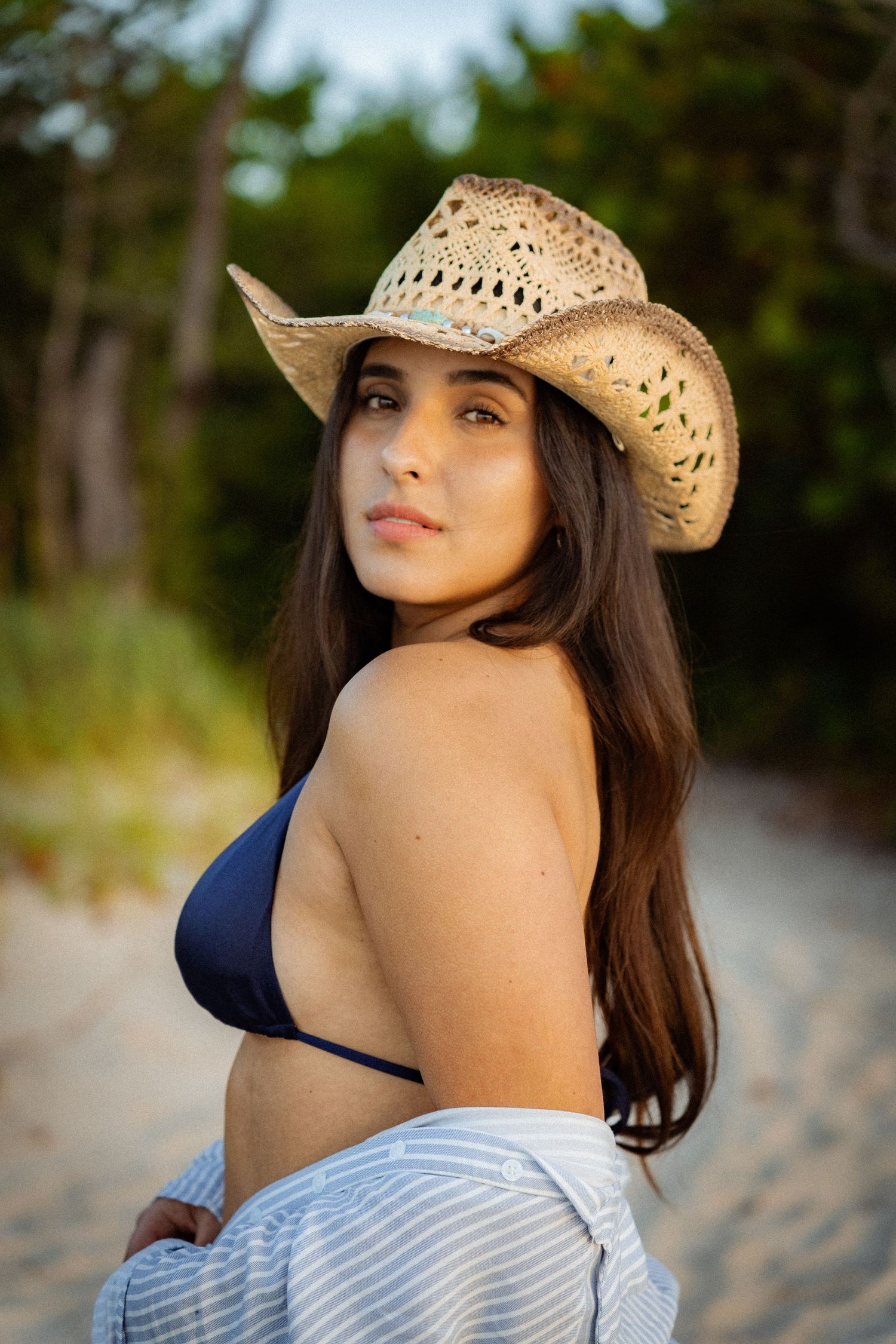 Sabrina Palma. Woman wearing a straw cowboy hat and navy swimsuit top poses on a sandy beach at sunset.