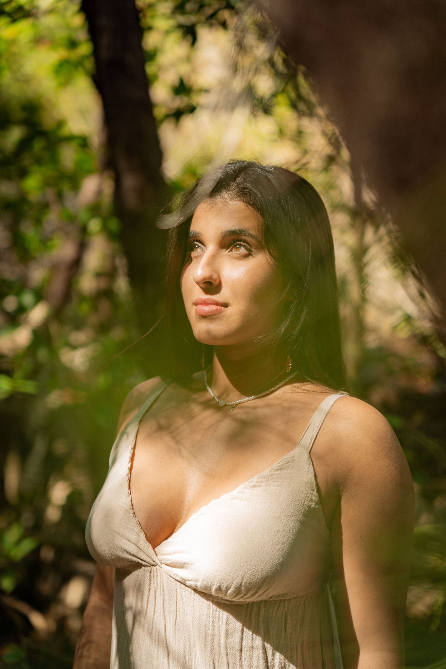Sabrina Palma. Woman wearing a white dress poses thoughtfully in a sunlit forest with green foliage in the background.