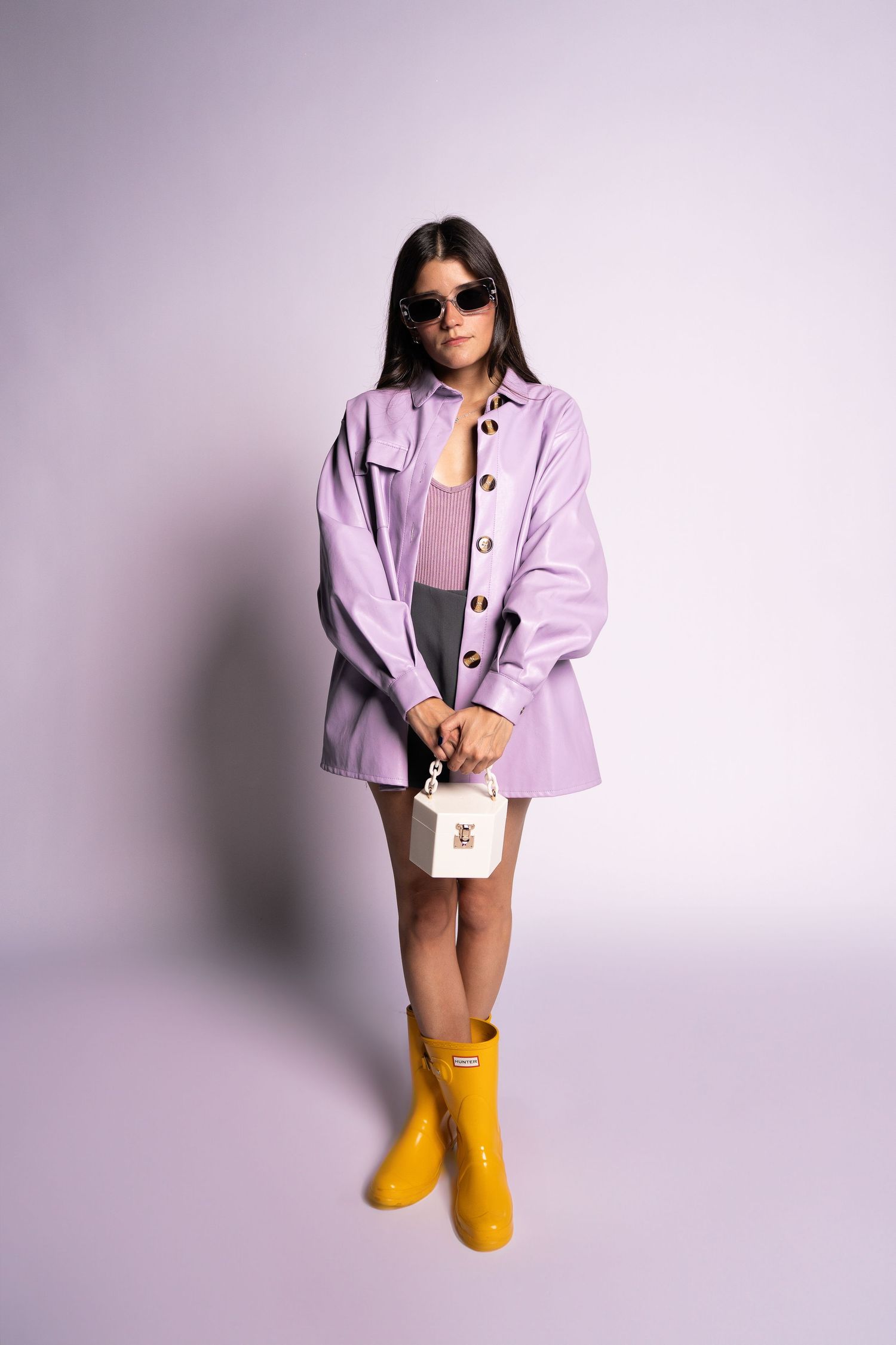 Sarah Sanchez. Sasainforma. Stylish fashion model wearing lilac jacket, sunglasses, and yellow rain boots against purple background.