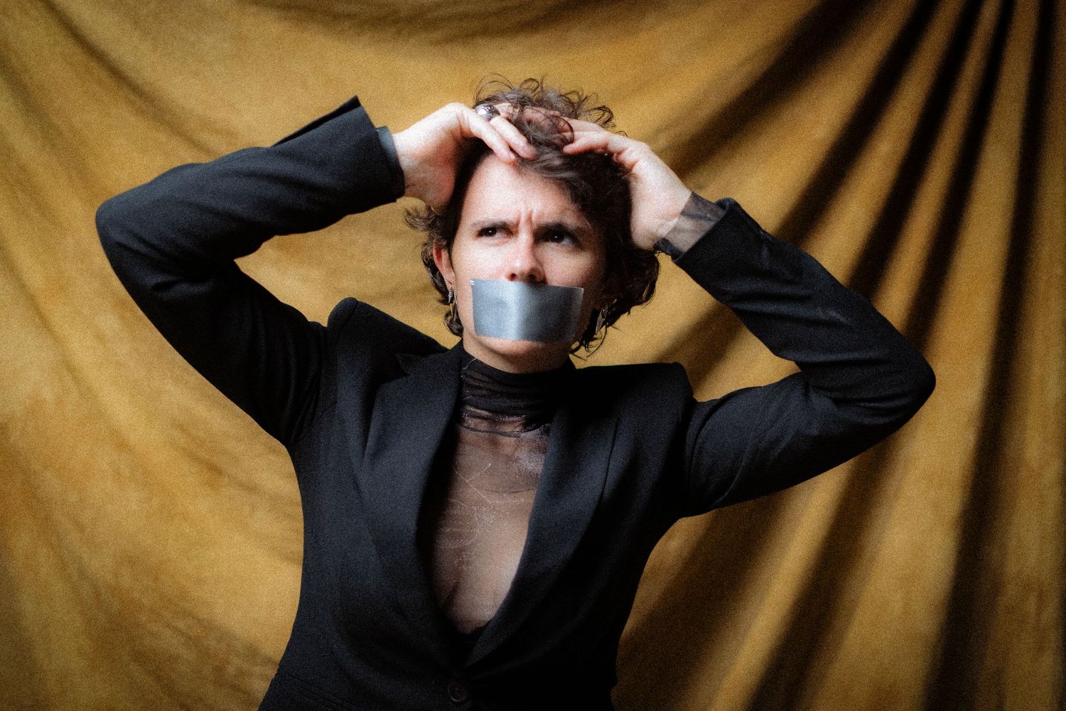 Isabel Römer. Artistic portrait against gold backdrop showing figure in black suit with duct tape across mouth.