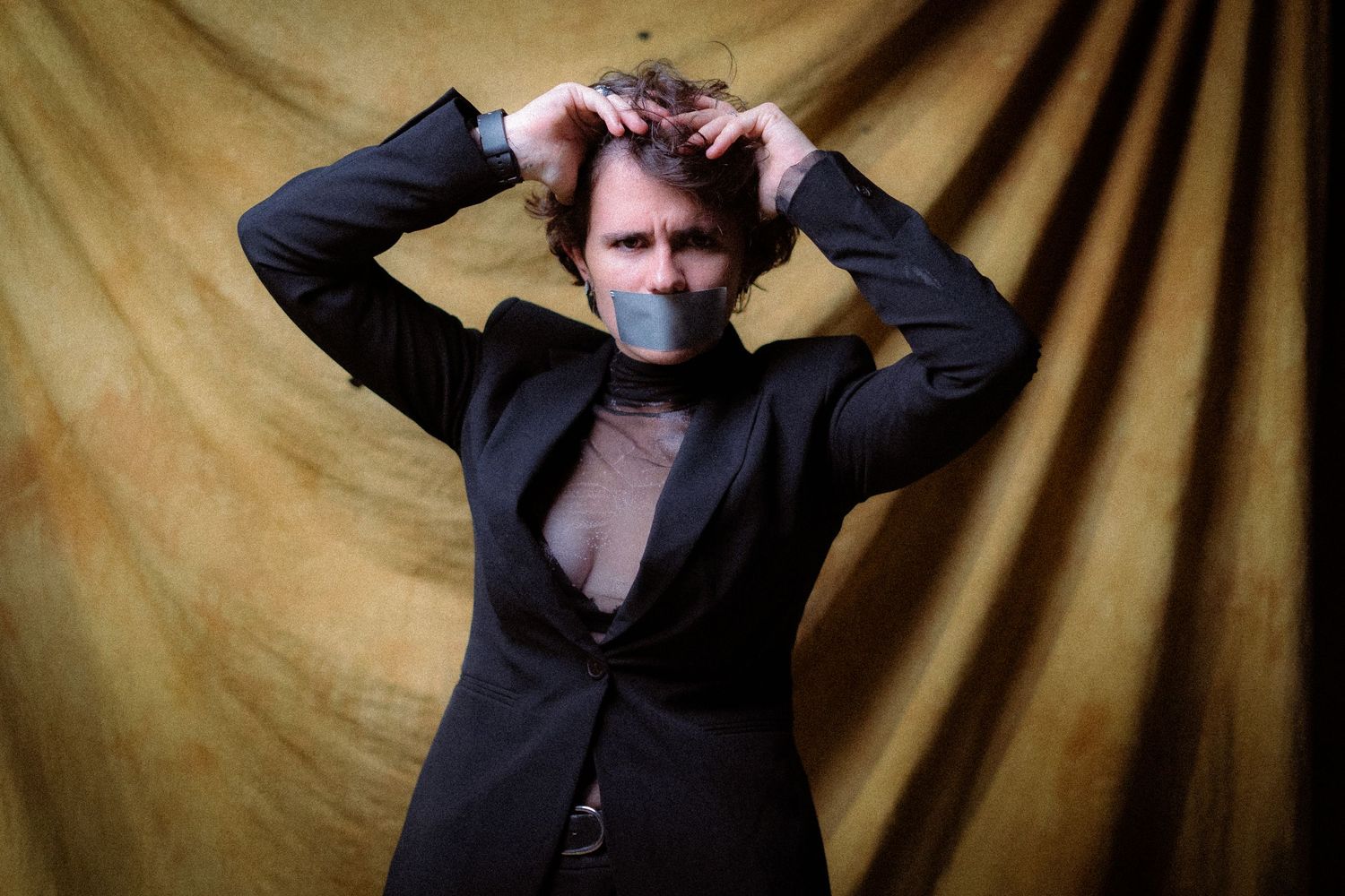 Isabel Römer. Moody portrait of person in black attire gesturing dramatically against golden backdrop.