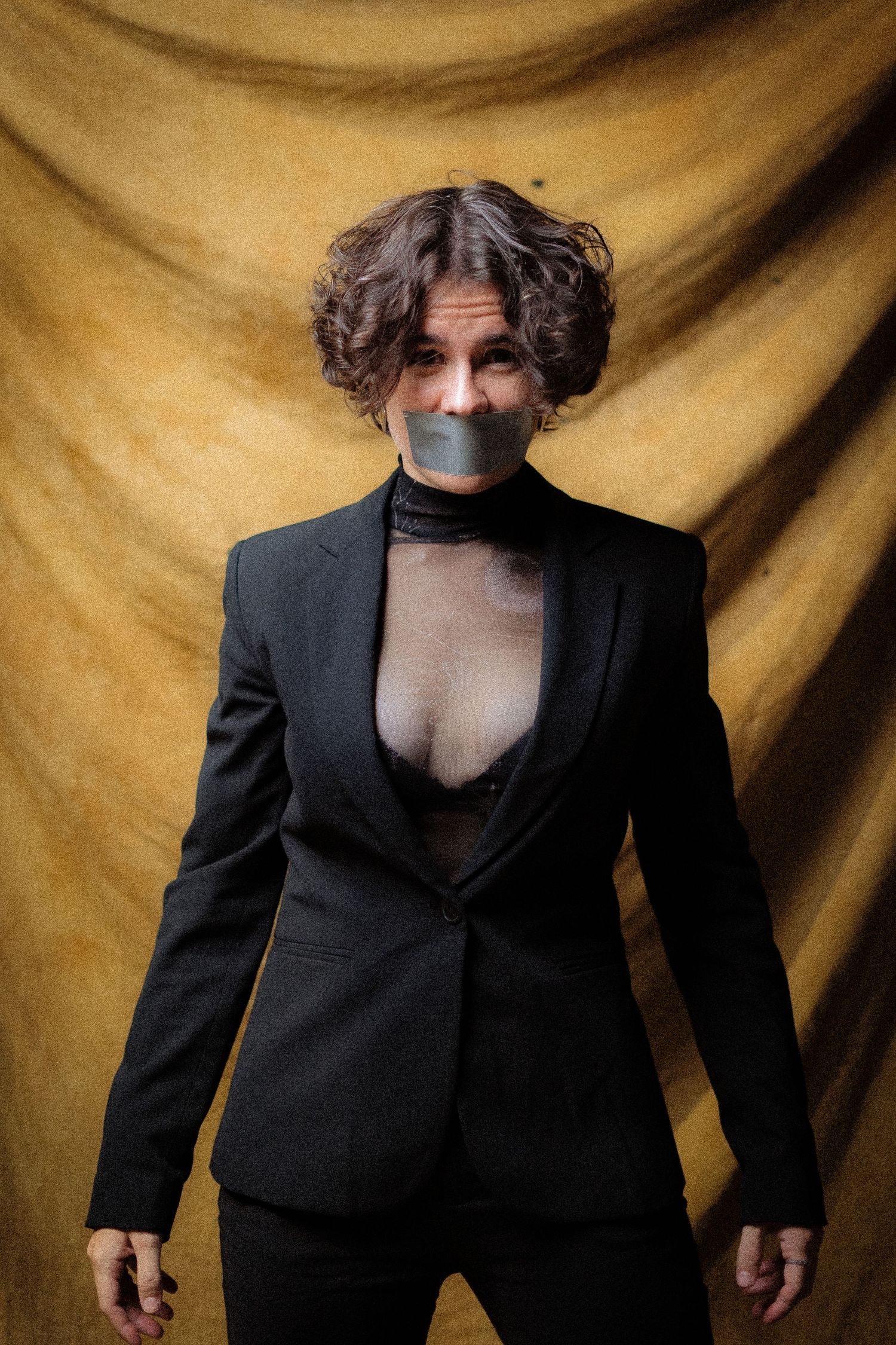 Isabel Römer. Dramatic portrait with person in black suit and tape across mouth against golden backdrop.