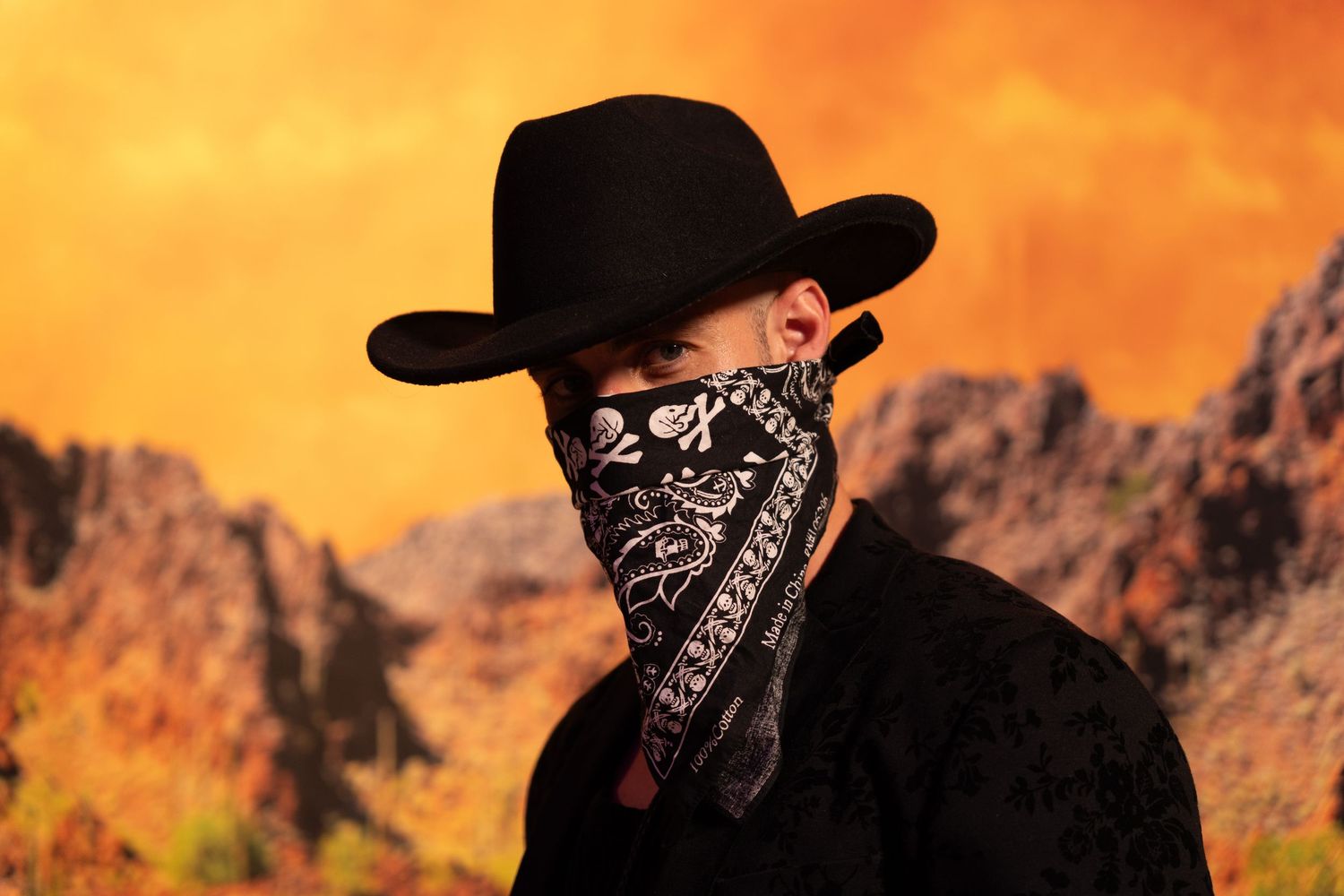 Silhouette against orange desert sunset wearing black cowboy hat and bandana with rocky mountains in background.