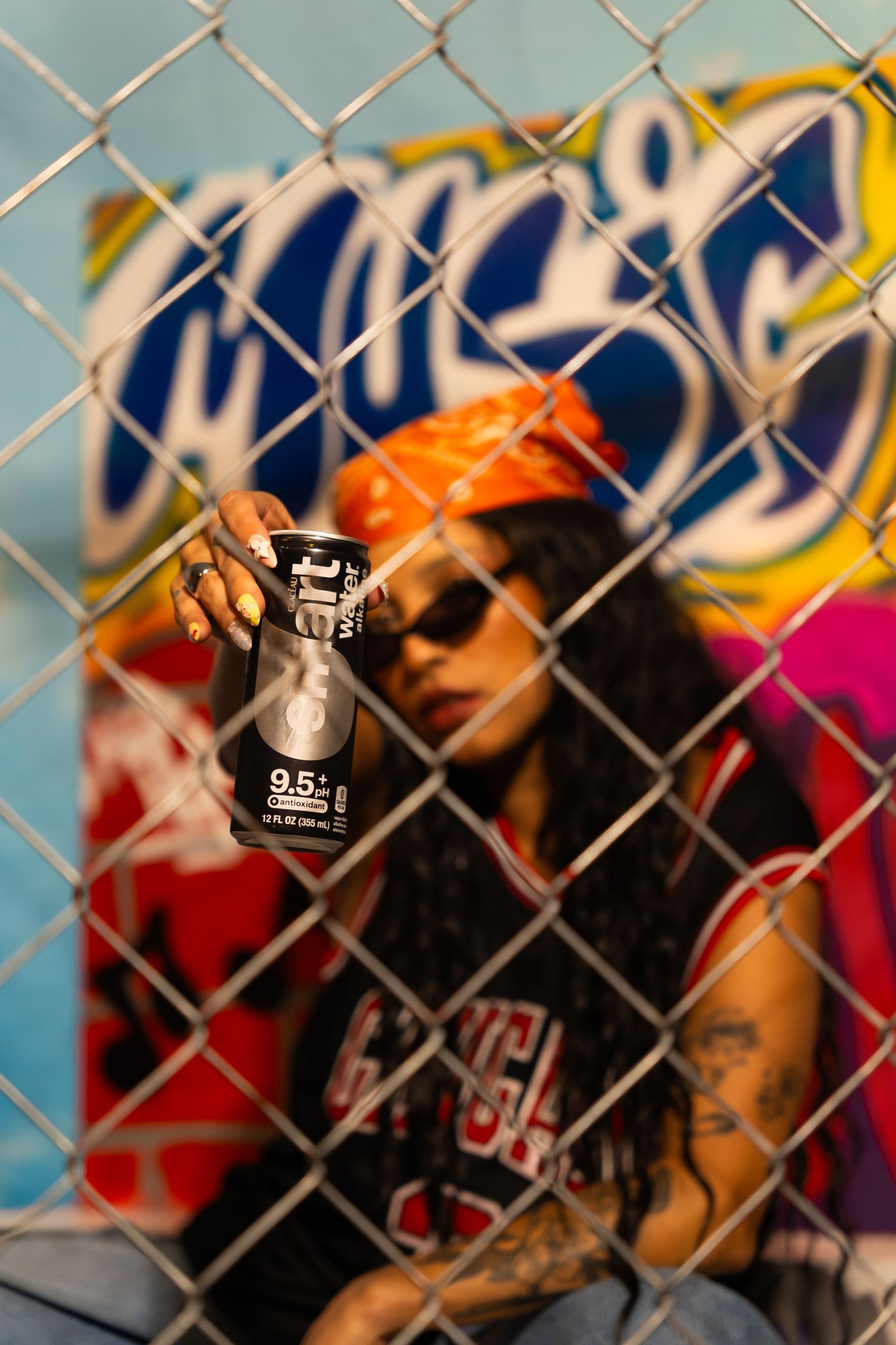 Through a chain link fence, someone holds up a black can of Smart Water with graffiti art visible in the background.