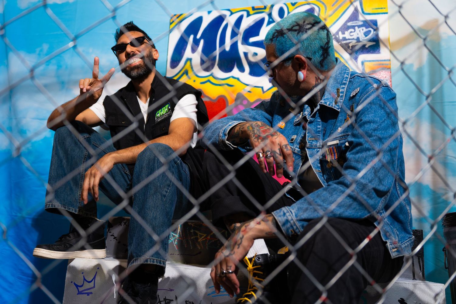 Two men in casual attire sit together against a chain-link fence with colorful graffiti art in the background.