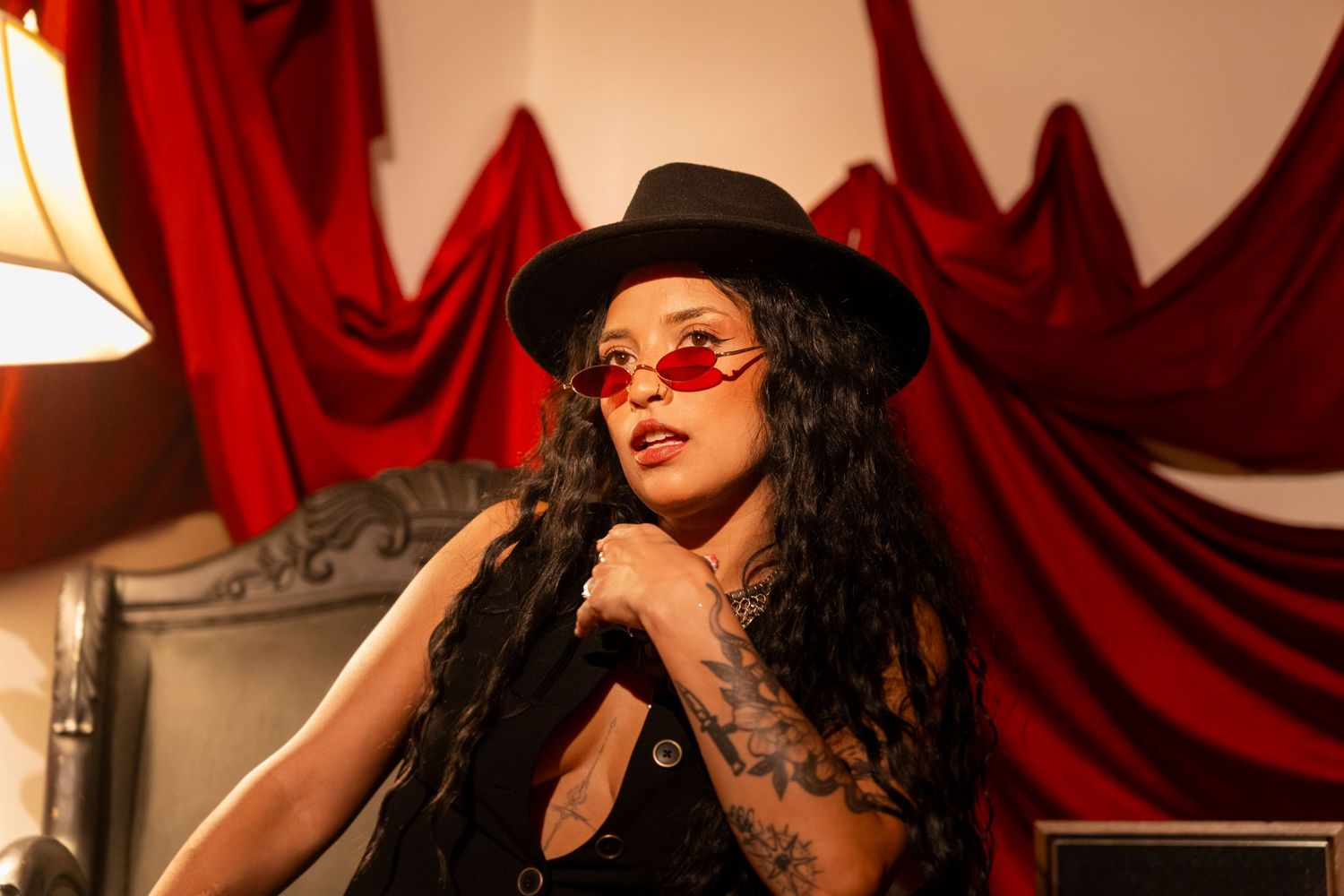 Figure in black attire with sunglasses and hat poses against red velvet curtain backdrop in dramatic lighting.