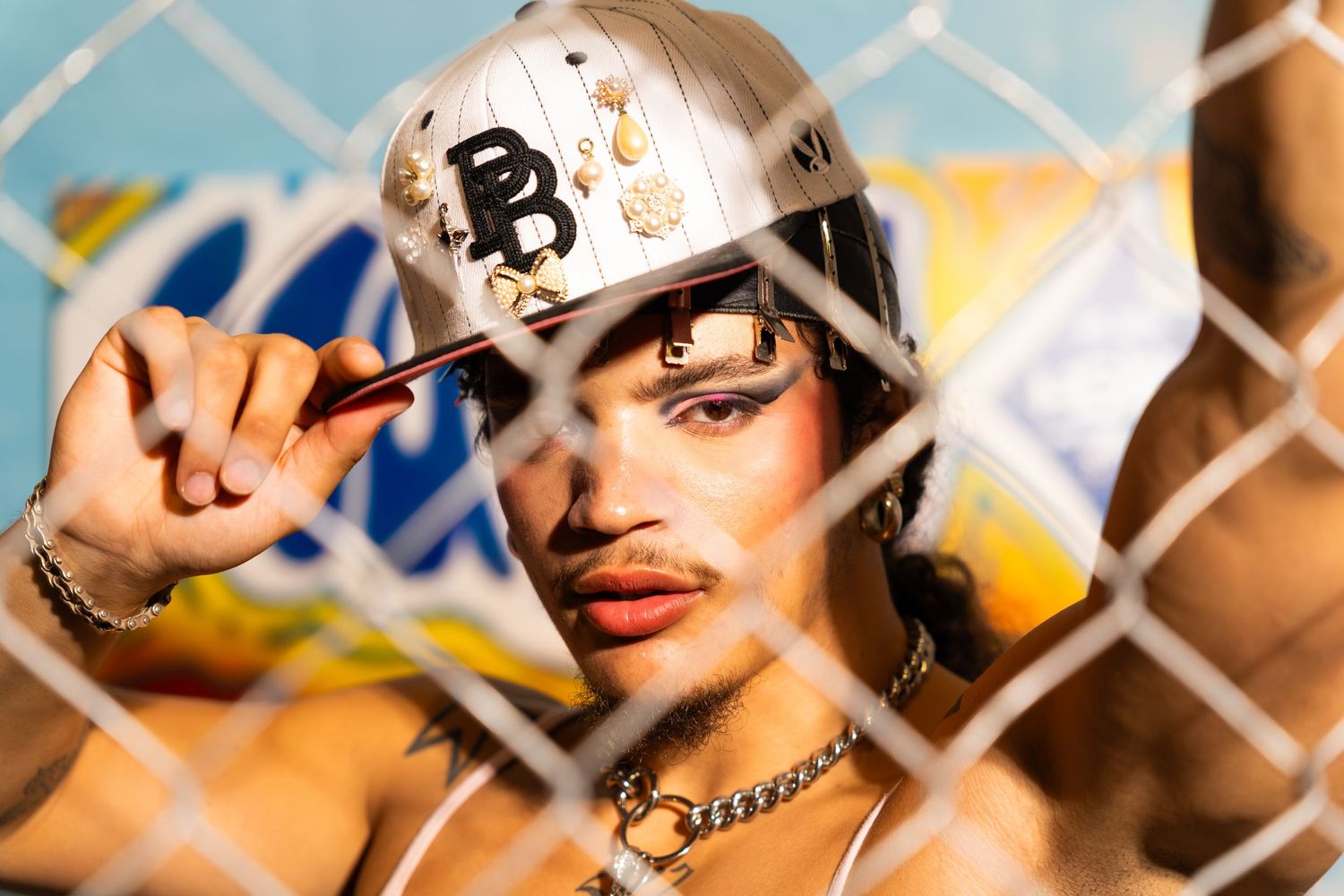 A person wearing a white baseball cap with 'B' logo poses behind a chain link fence in dramatic lighting.
