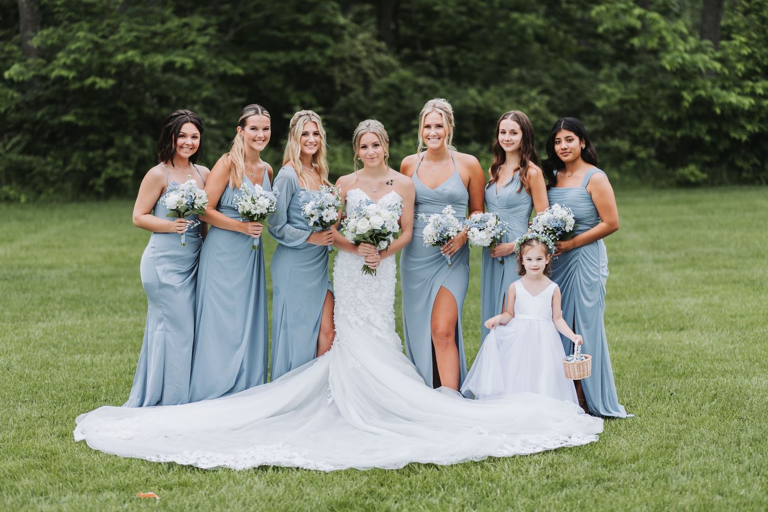 A bride in white wedding dress poses with bridesmaids wearing light blue gowns on a grass lawn at an outdoor wedding.