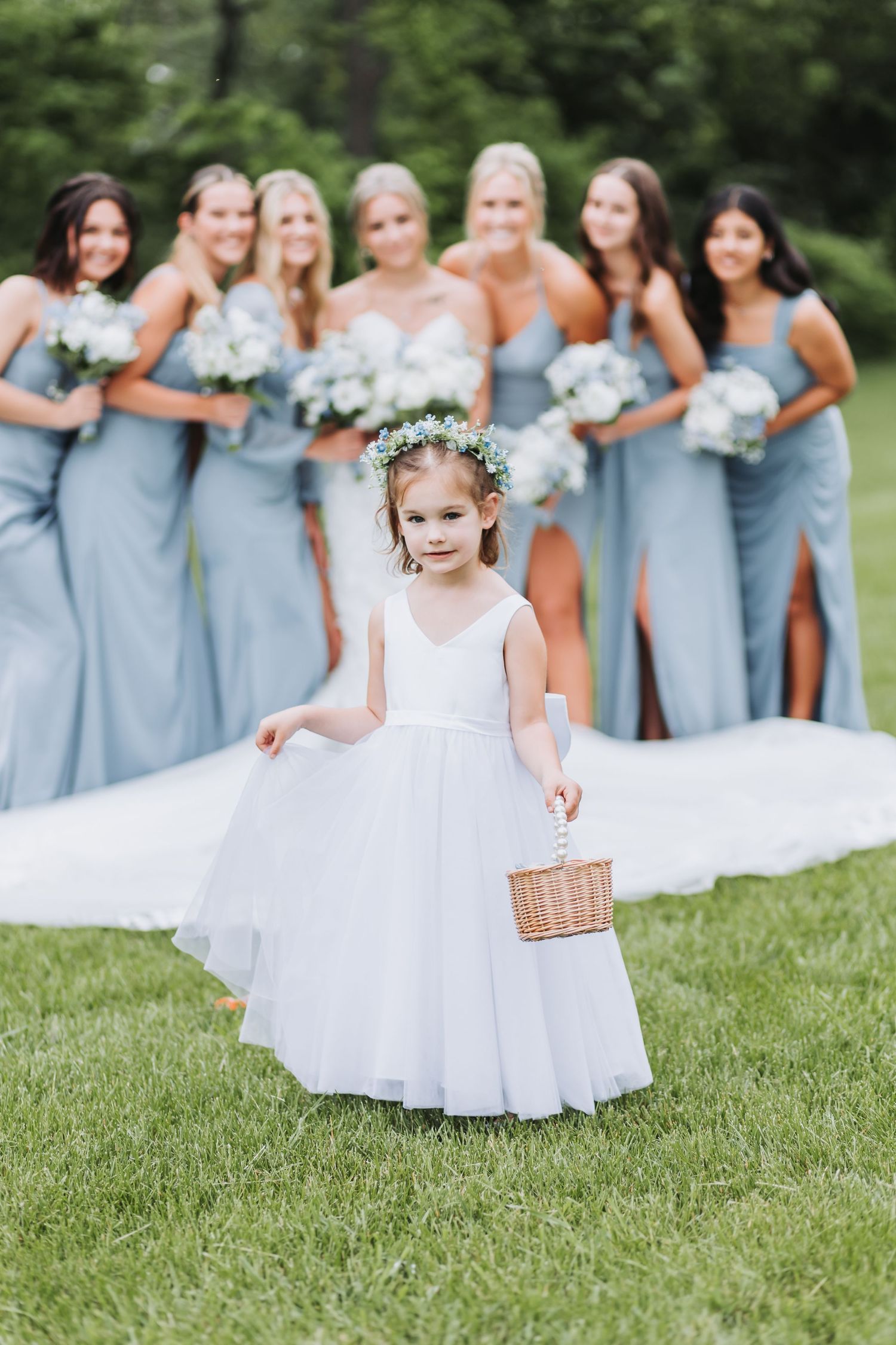 Flower girl in white dress holds basket while bridesmaids in light blue dresses stand behind on grassy lawn.