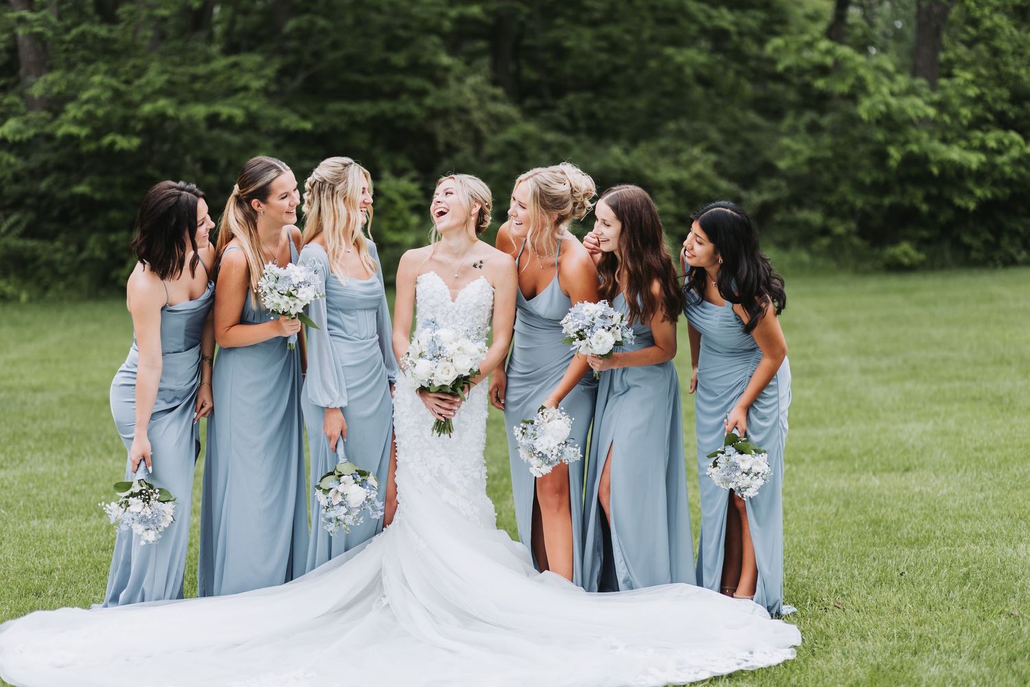 Bridesmaids in light blue dresses gather on a green lawn with a bride wearing a flowing white wedding gown.