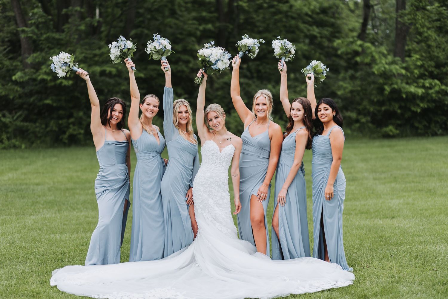 A bridal party in powder blue dresses and white bouquets poses on grassy lawn during summer wedding photoshoot.