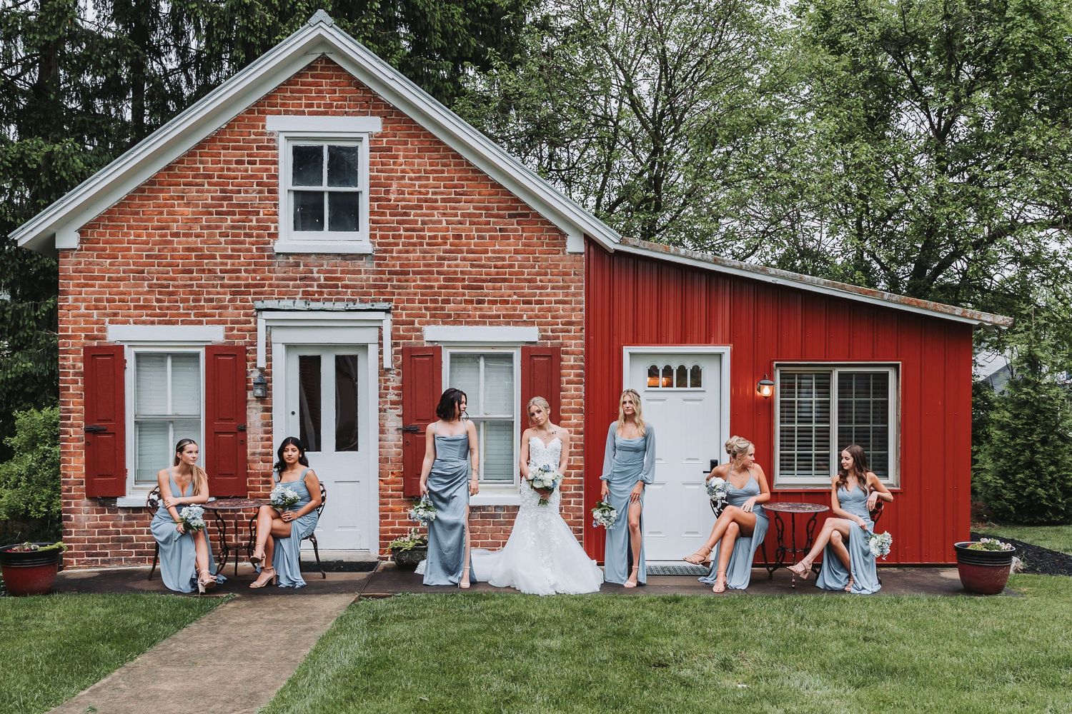 A rustic red brick and wooden barn serves as a charming backdrop for a wedding party lounging outside.