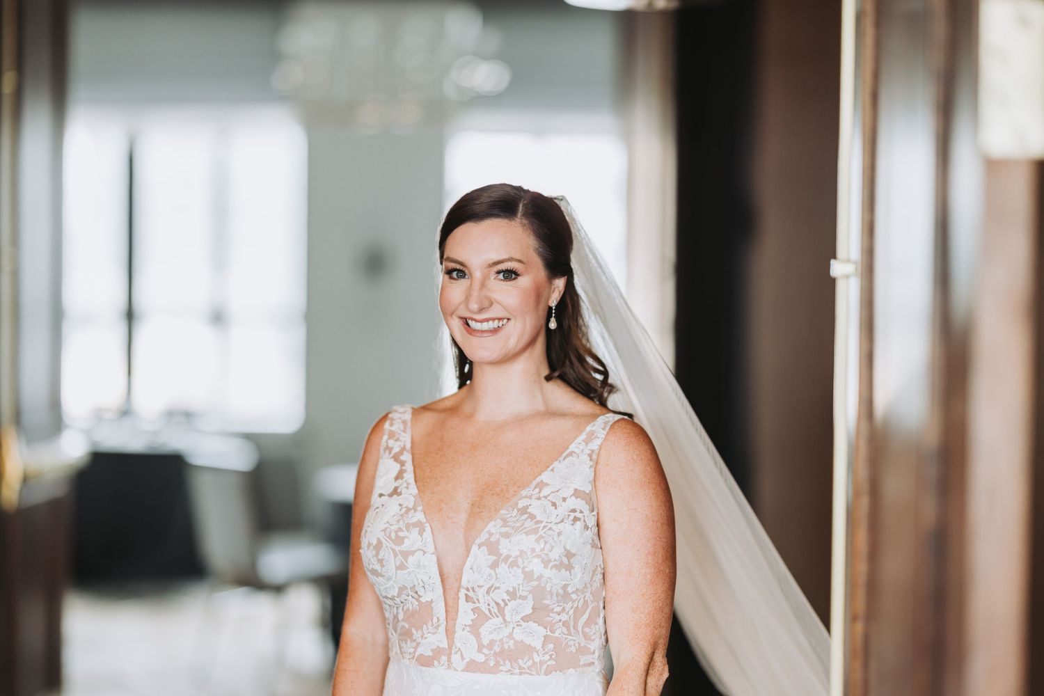 A bride smiles while wearing a deep v-neck lace wedding gown with a flowing veil.