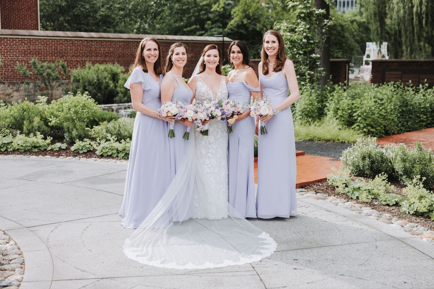 A bridal party poses outdoors wearing light purple bridesmaid dresses and holding floral bouquets.
