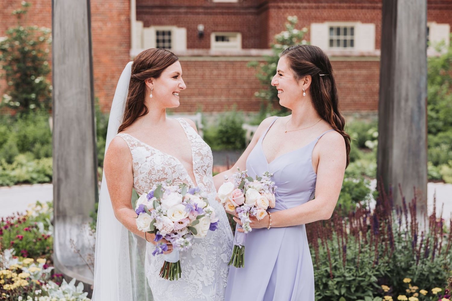 Two people wearing lavender and white gowns stand together holding pastel floral bouquets in a garden setting.
