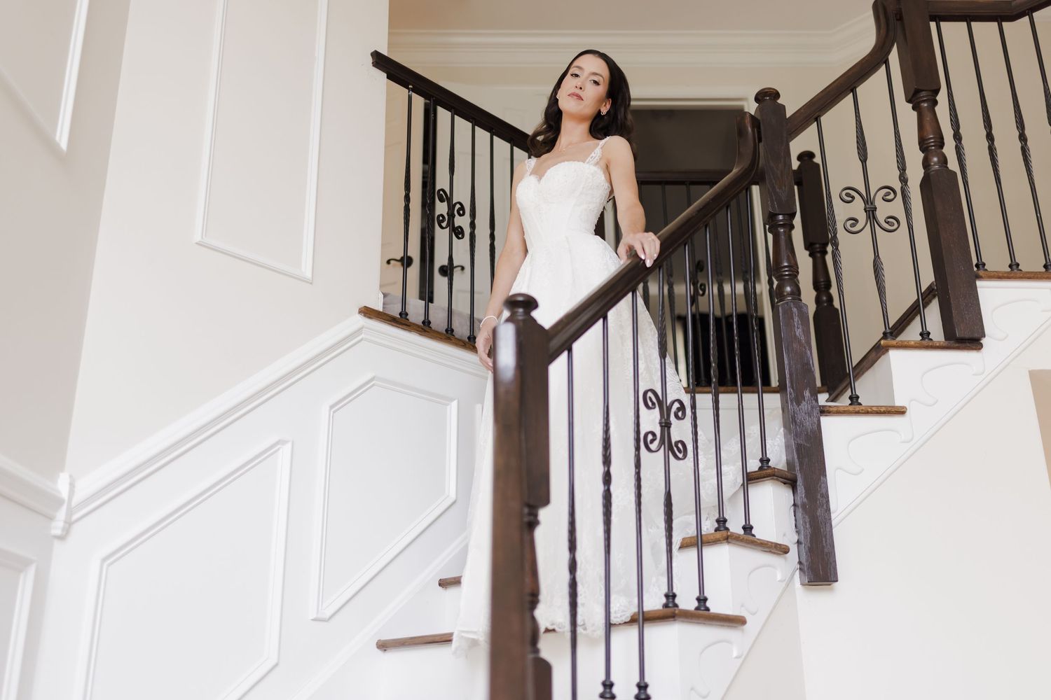 Elegant bride in a white strapless wedding dress poses gracefully on a grand wooden staircase with wrought iron railings.
